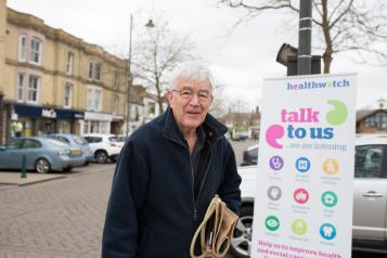 Elderly man standing in front of a Healthwatch banner saying "Talk to us"